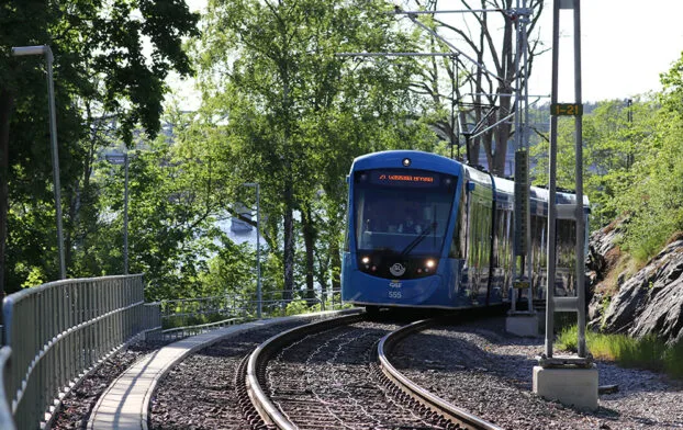 Blå spårvagn i kurva vid vatten och lövträd